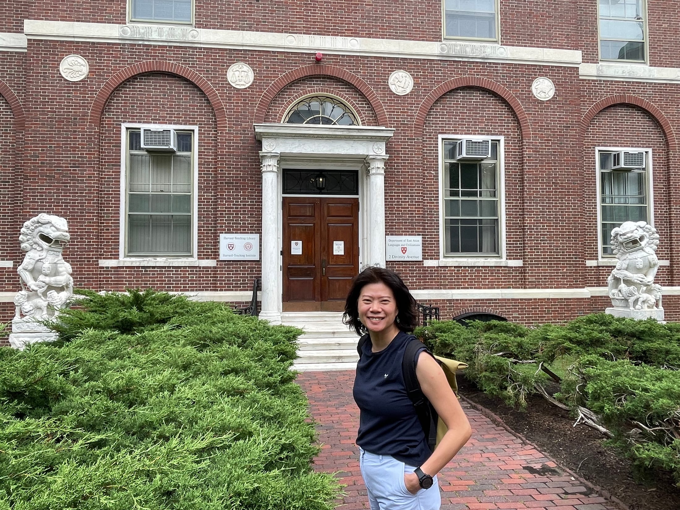 Jiraporn Laocharoenwong outside the Harvard-Yenching Institute, a red-brick building with a white columned doorway flanked by two stone guardian lions.